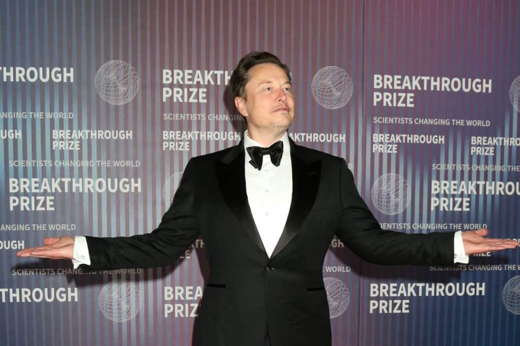 Elon Musk com o rosto de lado e os braços abertos