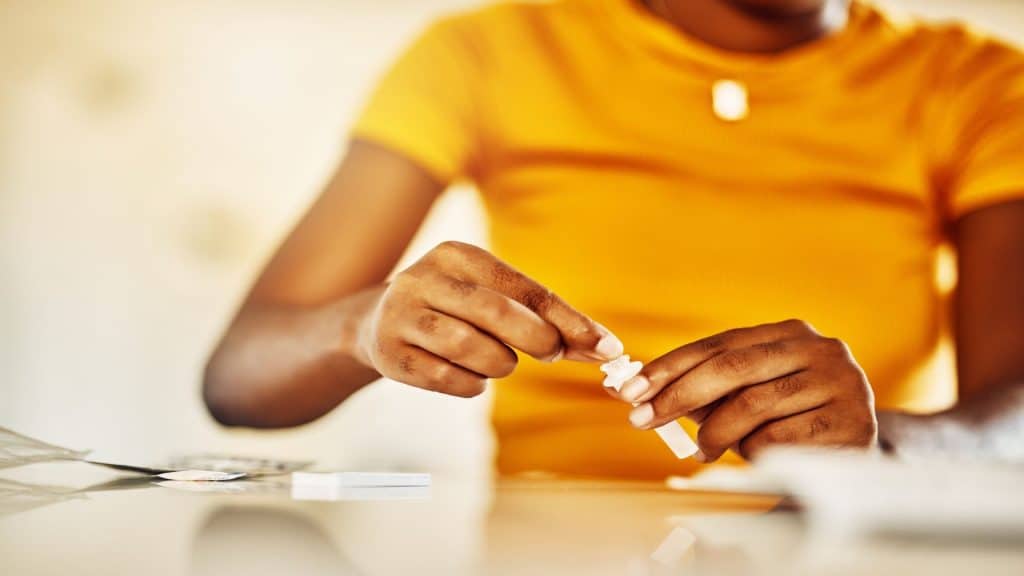 Mãos de uma mulher africana com HIV e AIDS em casa com um kit de teste de sangue sentada à mesa esperando para verificar os resultados. Close-up de uma jovem afro com remédios ou pílulas para uma condição médica.