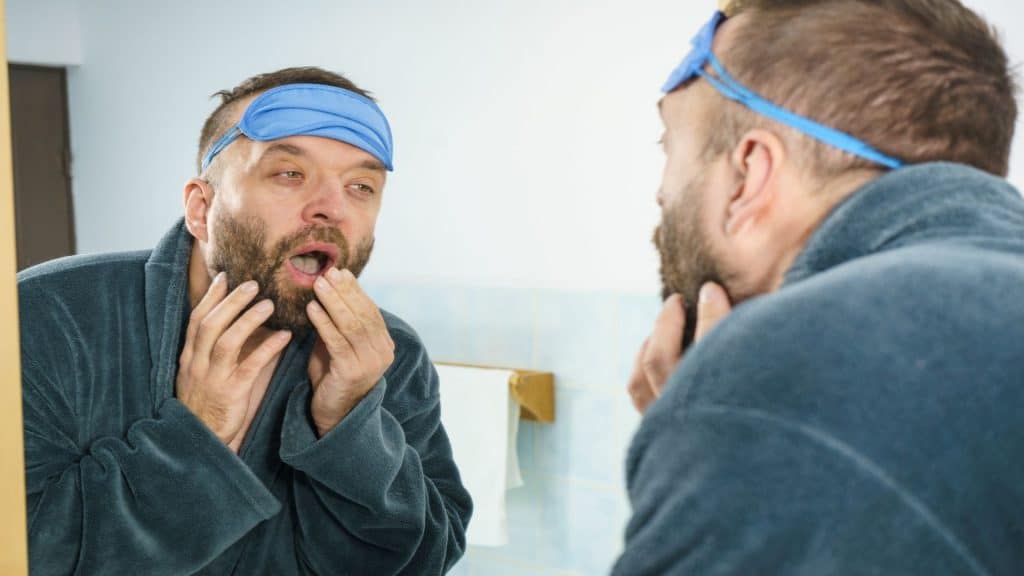 Homem adulto engraçado com máscara de dormir na testa tendo dificuldades para acordar. Em pé na frente do espelho olhando para seu rosto, bocejando, parecendo cansado e com sono/Shutterstock - Foto Colaborador Voyagerix