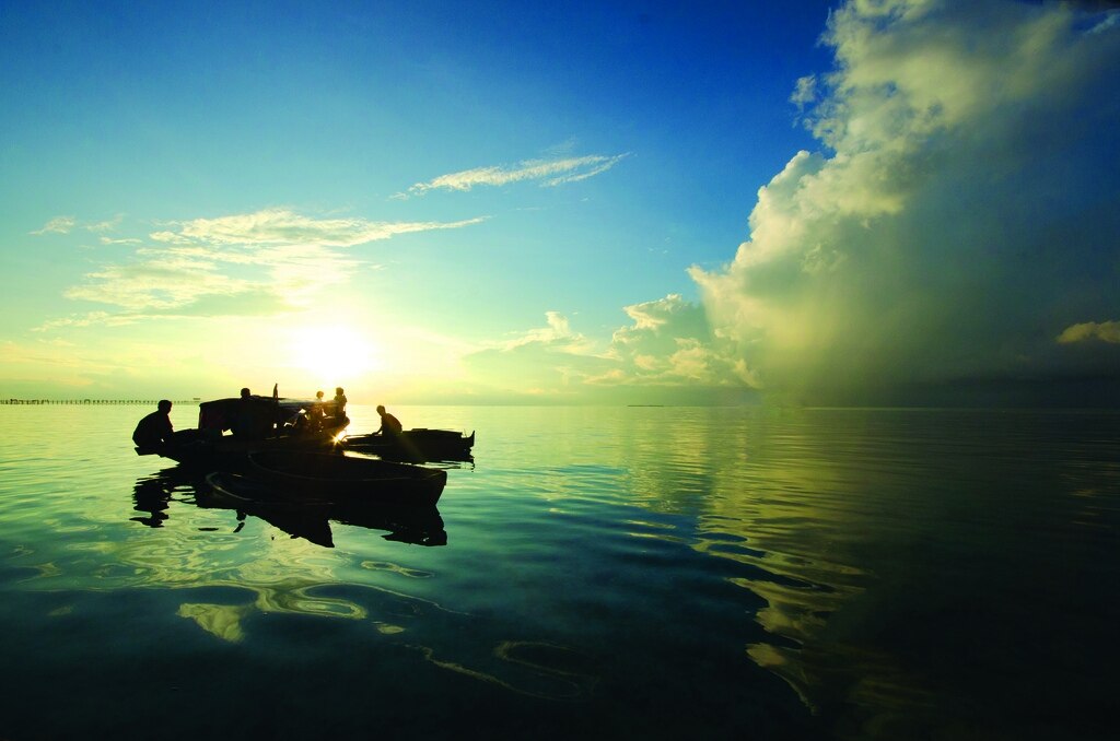 Silhuetas de pessoas Bajau em pequenos barcos tradicionais sobre águas calmas durante o pôr do sol, com o céu azul misturado a tons de amarelo e nuvens ao fundo.