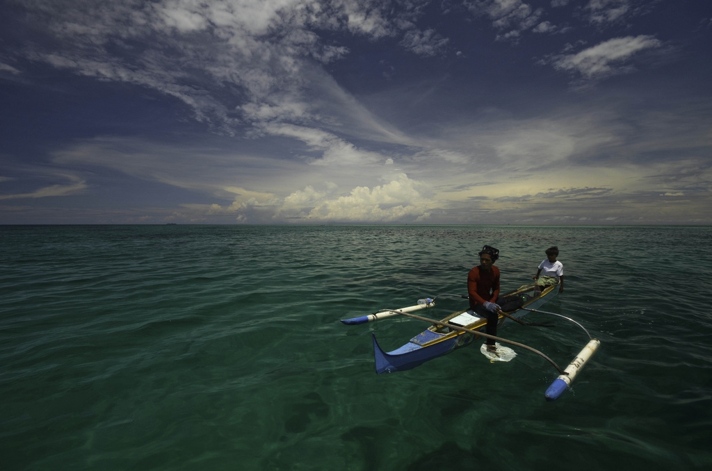 Dois homens Bajau sentados em um barco tradicional de casco duplo, feito de madeira, flutuando sobre águas cristalinas de cor esmeralda. O céu ao fundo apresenta nuvens brancas esparsas e uma tonalidade azul vibrante, criando uma paisagem marítima serena.