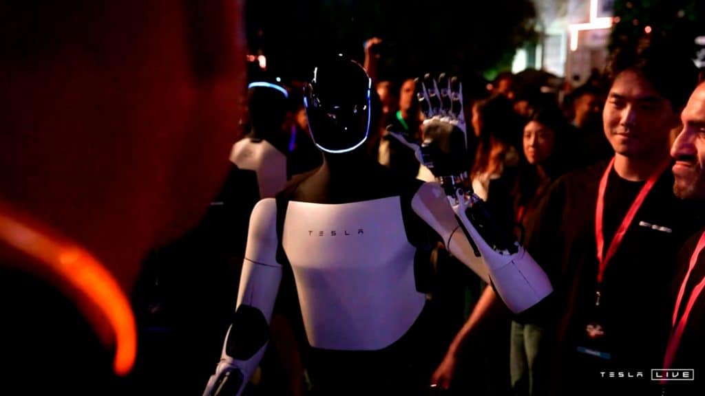 Robô Optimus acenando rodeado de pessoas durante evento da Tesla