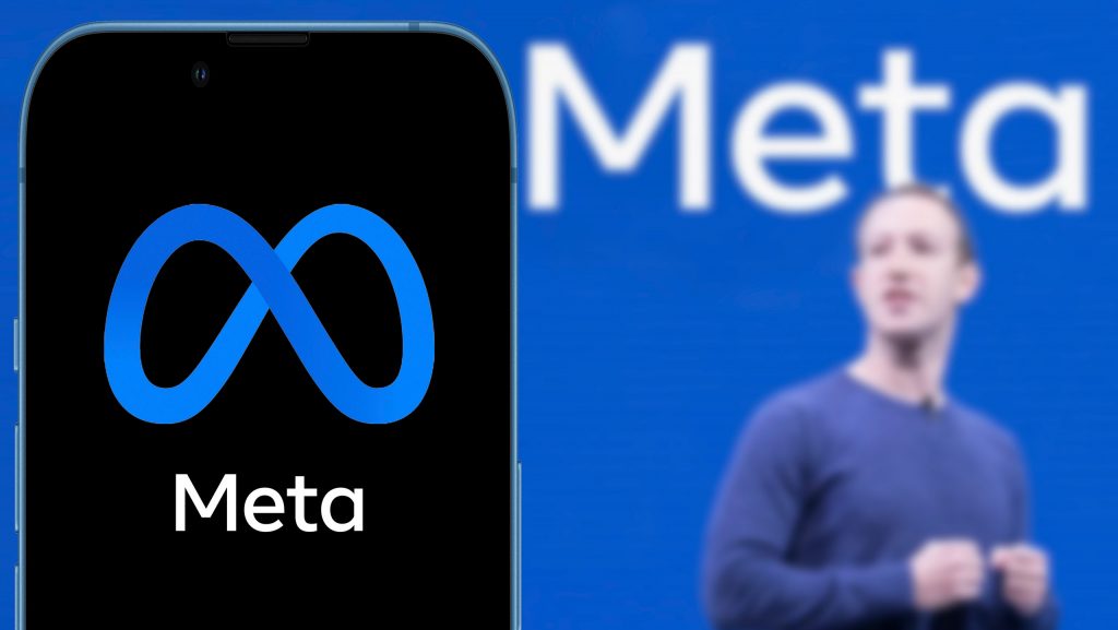 Smartphone com logo da Meta; ao fundo, desfocado, placa azul com o escrito "Meta" e, mais adiante, Mark Zuckerberg