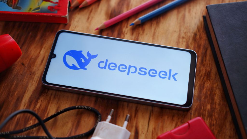 Celular com logotipo do DeepSeek na tela colocado sobre mesa de madeira rodeado de materiais de escritório como lápis e carregador