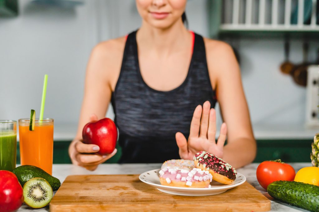 Imagem mostra mulher querendo parar de comer açúcar ao segurar uma maçã em uma das mãos e fazer gestro com a outra para resistir a um prato de doces