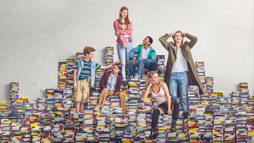 Imagem promocional da série 'Everything Sucks!'. Seis adolescentes posam sobre uma pilha de fitas VHS, formando uma escadaria. O fundo é um muro branco. Cada personagem tem um estilo distinto, refletindo a estética dos anos 90.