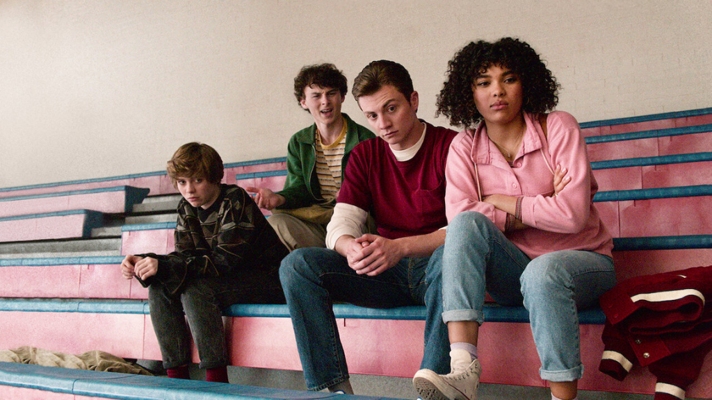 Arte promocional da série "I Am Not Okay with This" (2020), da Netflix. Quatro jovens estão sentados em arquibancadas de ginásio, com expressões sérias e pensativas. A personagem à esquerda veste um suéter escuro e parece preocupada, enquanto os outros três olham em direções diferentes, transmitindo um clima de tensão e incerteza.