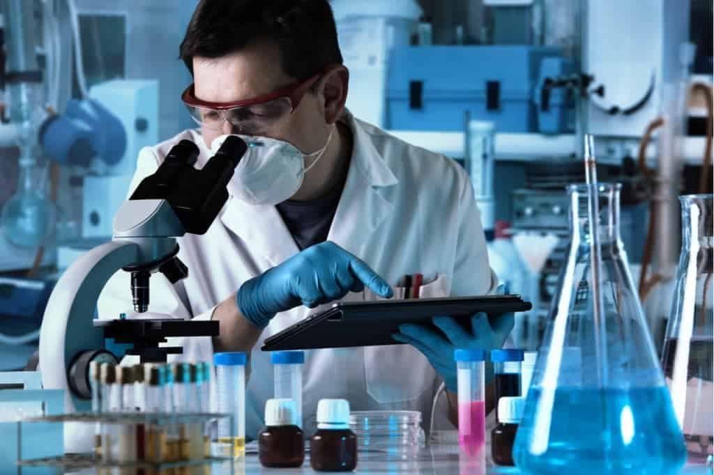 Imagem mostra um cientista observando experimentos no microscópio, enquanto manipula um tablet