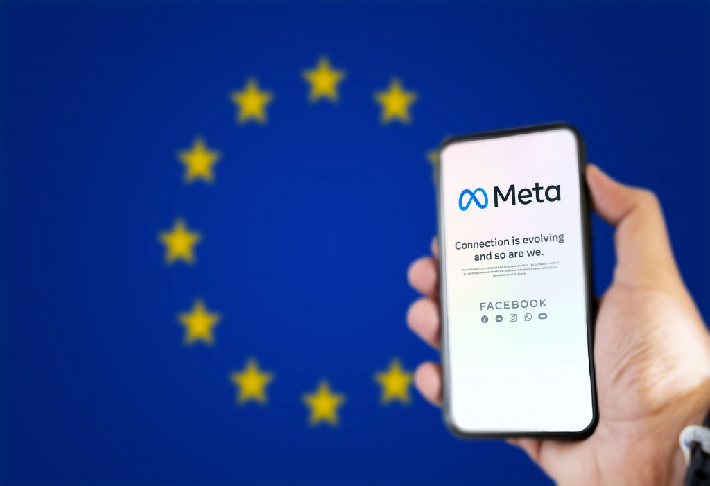 Ao fundo, desfocada, a bandeira da União Europeia; à direita, pessoa segurando um smartphone, no qual é exibida uma página da Meta