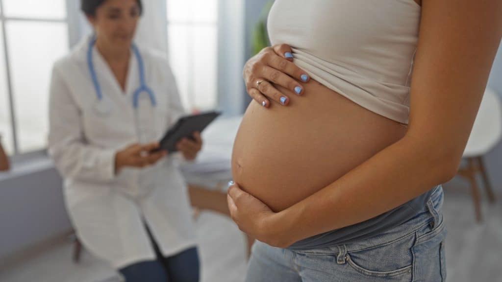 Mulher grávida com barriga em uma sala de clínica enquanto o médico se senta com um comprimido fornecendo consulta médica