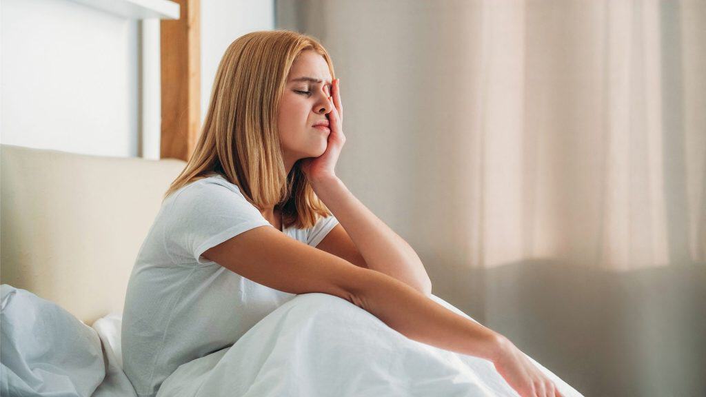 Insônia, cansaço. Dor de cabeça, fadiga. Depressão matinal. Desespero, jovem garota fraca esfregando o rosto, franzindo a testa, sentindo-se mal de manhã cedo em quarto branco com espaço livre.