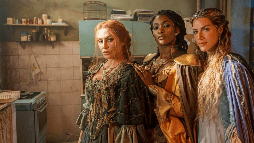 Três mulheres vestindo trajes medievais e místicos posam em uma cozinha simples e desgastada