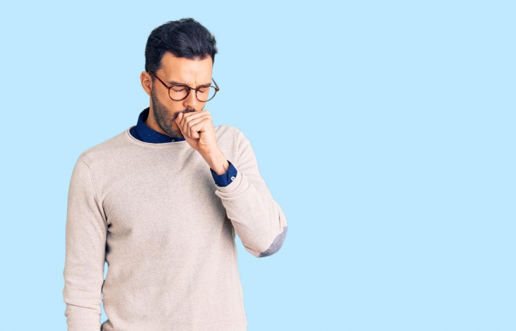 homem de blusa de frio e óculos com um fundo azul atrás, tossindo com a mão na boca