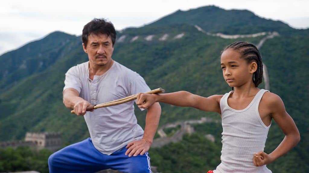 Homem ensinando artes marciais a um menino em um cenário ao ar livre, com montanhas verdes e a Grande Muralha ao fundo