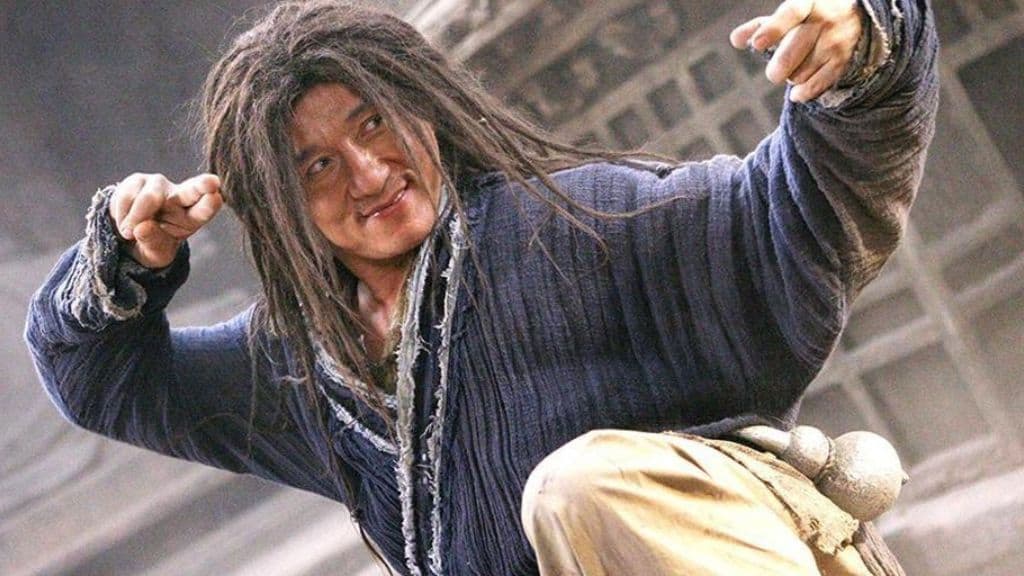Homem com cabelos desgrenhados, vestindo trajes rústicos e expressão determinada, em pose de combate em um cenário antigo