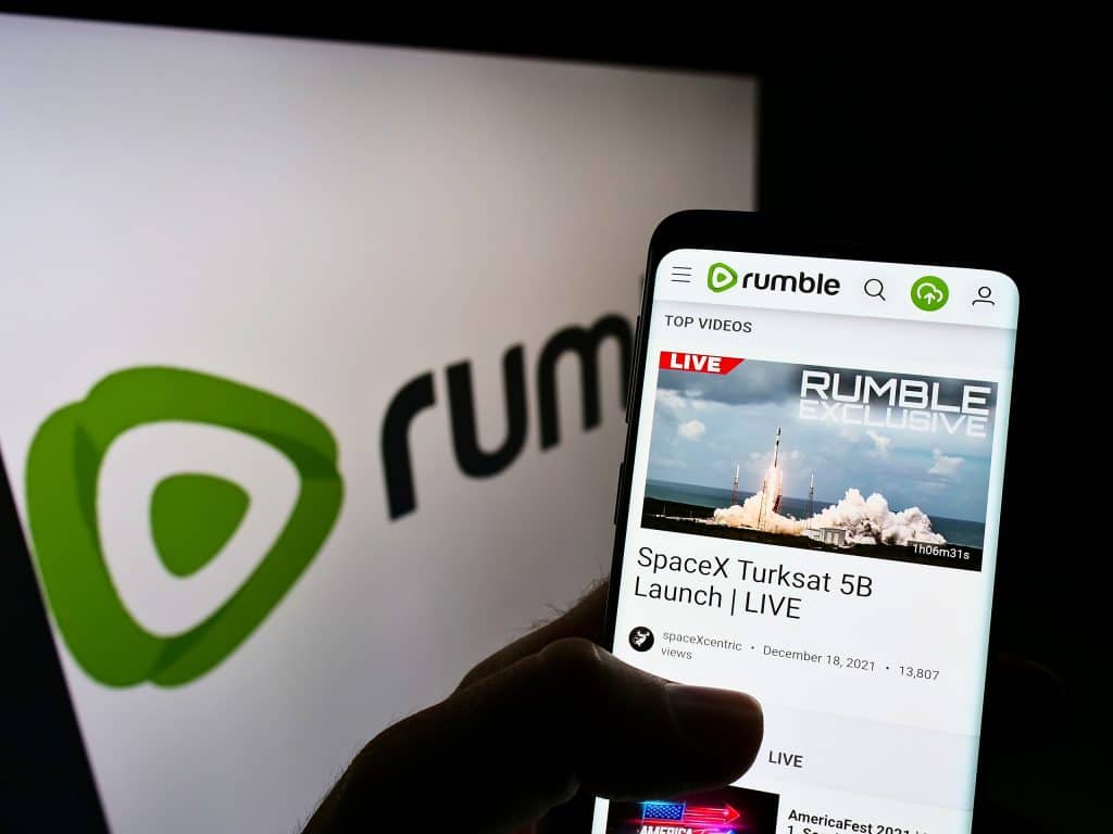 Ao fundo, logo do Rumble em um monitor; à frente, app da plataforma aberta em um smartphone
