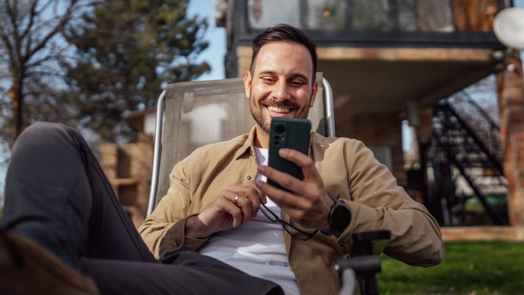 Um homem caucasiano com barba e óculos sentado ao ar livre em um dia ensolarado vestindo camisa, usando um smartphone para mensagens SMS, navegando na internet ou fazendo videochamadas, usando um aplicativo, com espaço para texto, pessoas reais.