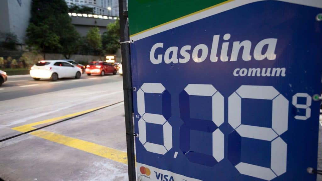 Foto de banner de posto de gasolina com preço de 6,79 a gasolina e ao fundo uma via expressa com carros parados no farol.