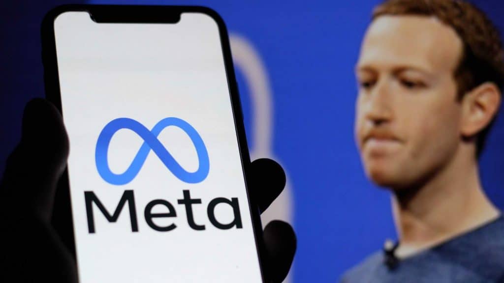 Celular com logomarca da Meta na tela na frente de monitor com foto de Mark Zuckerberg com cara franzida