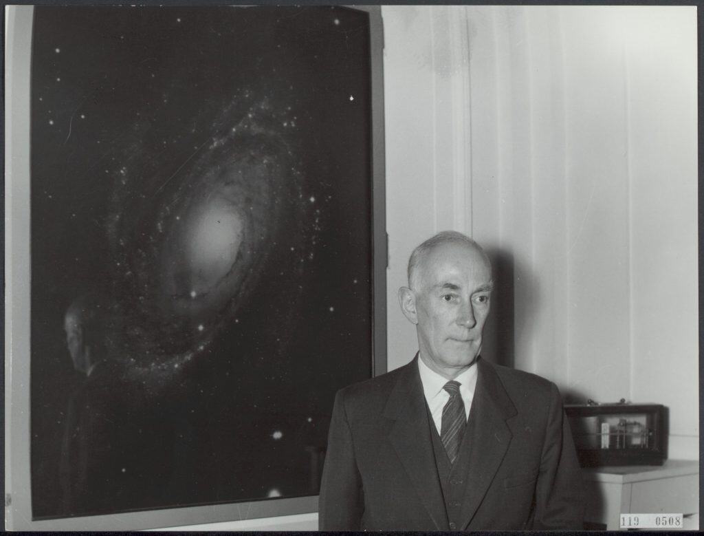 Retrato em preto e branco do astrônomo Jan Hendrik Oort ao lado de uma imagem de galáxia, em ambiente fechado.
