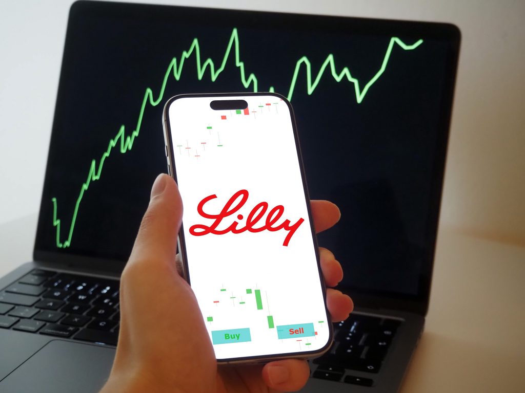 Logo da Lilly em um smartphone que está em uma mão; atrás, notebook com um gráfico