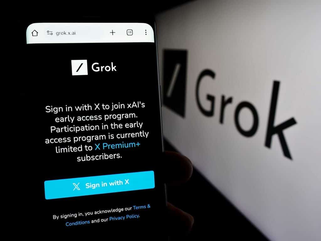 Página do Grok em um smartphone, ao lado do logo do chatbot impresso em uma parede