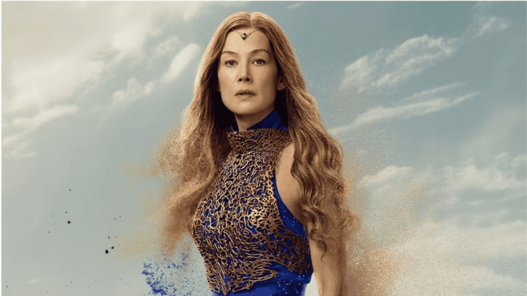 Rosamund Pike, caracterizada como Moiraine Damodred, veste um traje azul e dourado com detalhes intrincados. Seus longos cabelos ondulados esvoaçam ao vento, e um céu com nuvens serve de fundo. Partículas de poeira e tinta mágica flutuam ao seu redor