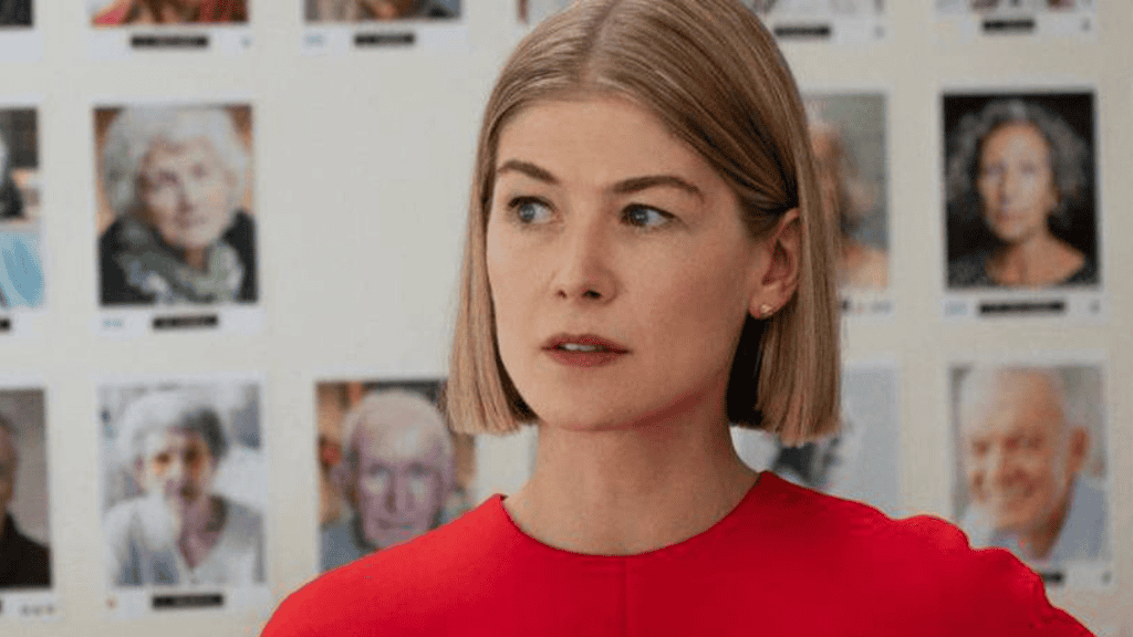 Rosamund Pike em cena de Eu Me Importo (2020), vestindo um vestido vermelho e exibindo uma expressão confiante. Ao fundo, uma parede coberta por fotos de idosos