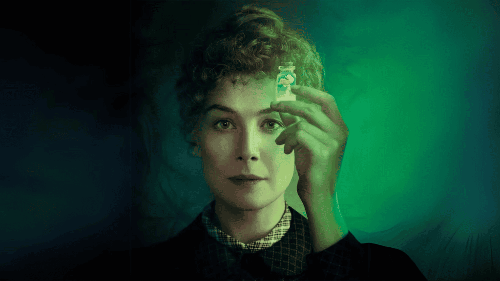 Rosamund Pike, caracterizada como Marie Curie, segura um pequeno frasco luminoso com uma substância radioativa, olhando atentamente para ele. O fundo apresenta tons esverdeados, evocando o brilho da radiação