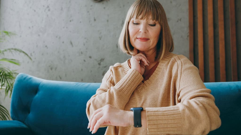 Idosa de 50 e poucos anos vestindo roupas casuais senta-se em um sofá azul, olha para um smartwatch, coloca a mão no pescoço para verificar o pulso, fica em casa em um apartamento para descansar, relaxar e passar o tempo livre na sala de estar interna com parede cinza.
