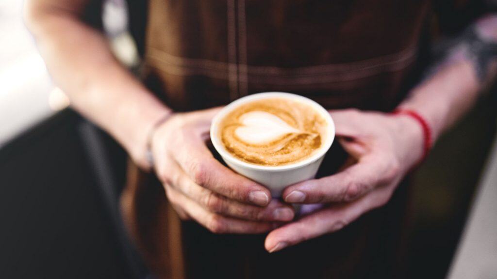 Barista segurando cappuccino, em pé no balcão. Barman de avental preparando bebida de café