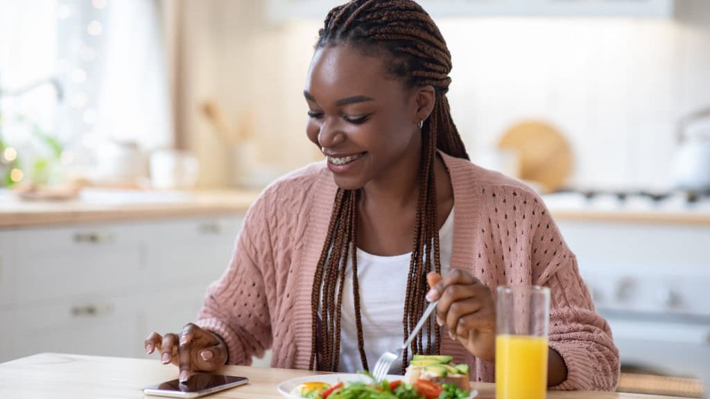 plicativo de contagem de calorias. Mulher negra sorrindo usando aplicativo moderno no smartphone enquanto toma café da manhã na cozinha em casa, senhora africana feliz sentada à mesa, verificando os dados diários de nutrição