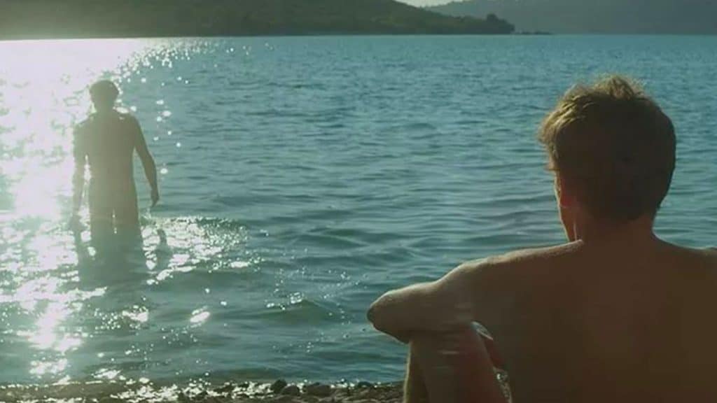 Homem sentado na areia olhando outro homem entrar na água em um lago.