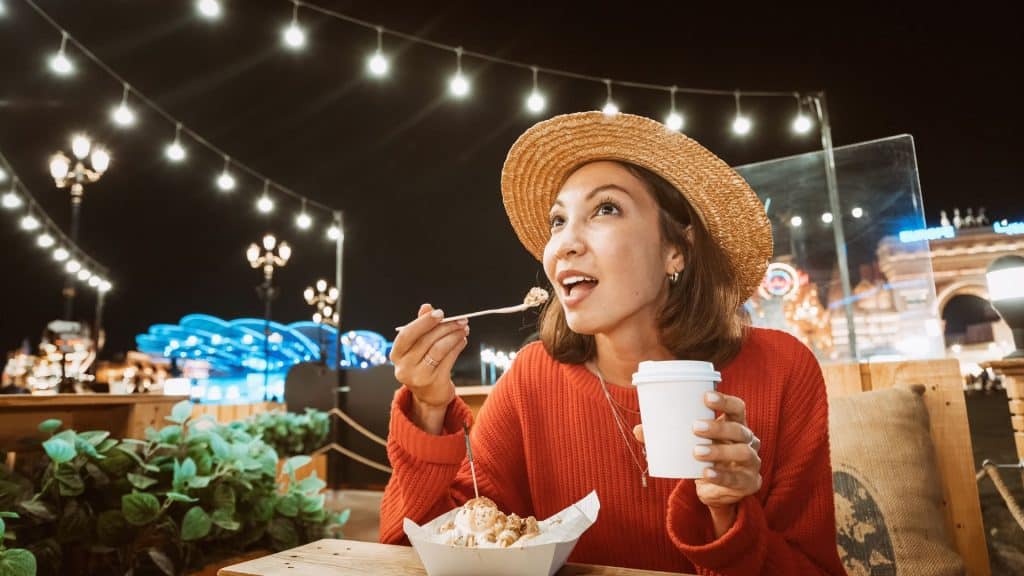 Mulher sorrindo come um bolo doce com sorvete e bebe café em um café noturno com luzes.