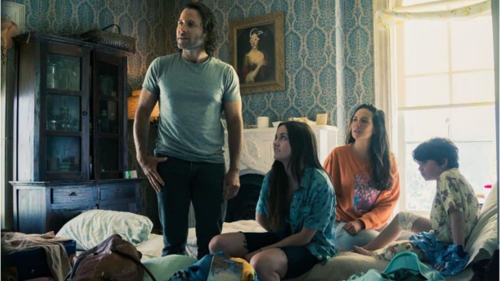 A imagem apresenta um quarto com decoração vintage. Há quatro pessoas, sendo uma em pé e três sentadas numa cama. O espaço inclui um papel de parede azul e branco, uma janela grande, um armário de madeira e itens espalhados, como malas e roupas.