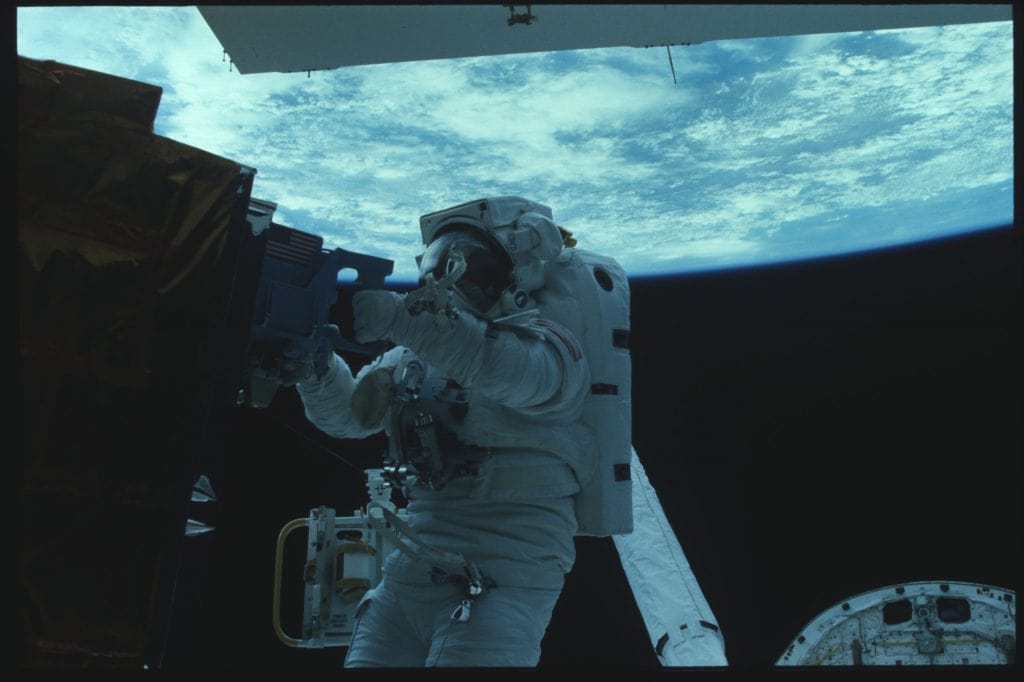 Astronauta James van Hoften durante caminhada espacial, reparando um satélite em órbita terrestre durante a missão STS-41C. A imagem mostra claramente o astronauta trabalhando com ferramentas específicas, tendo como fundo a curvatura azul da Terra vista do espaço.