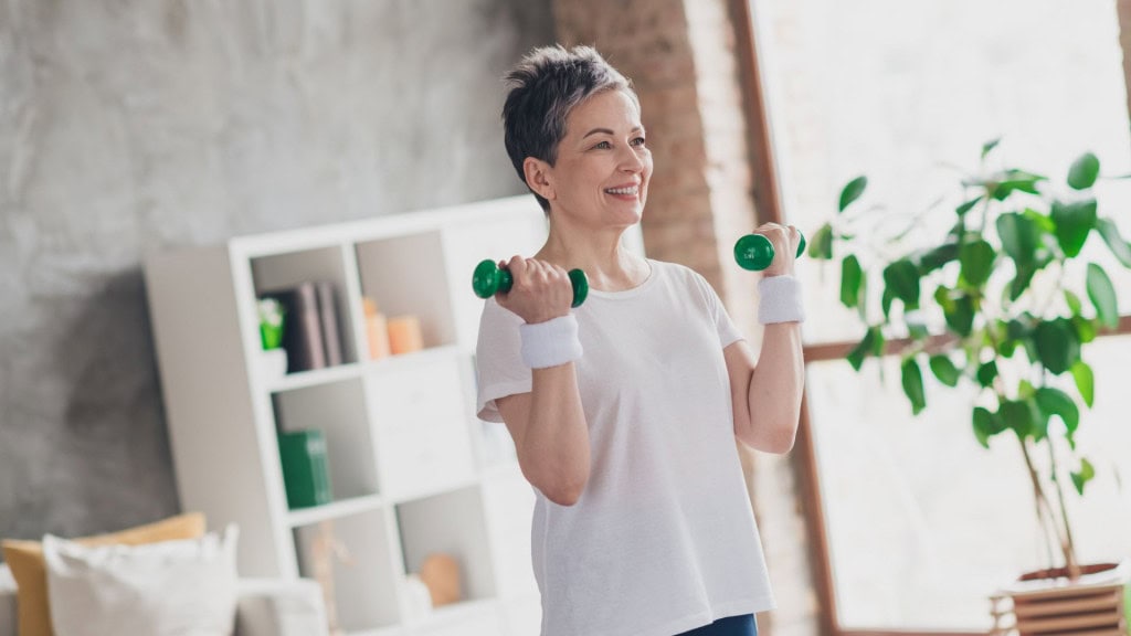 Foto de uma mulher idosa atraente fazendo exercícios com halteres em roupa esportiva em casa, em uma sala de estar espaçosa e iluminada, conceito de esporte.