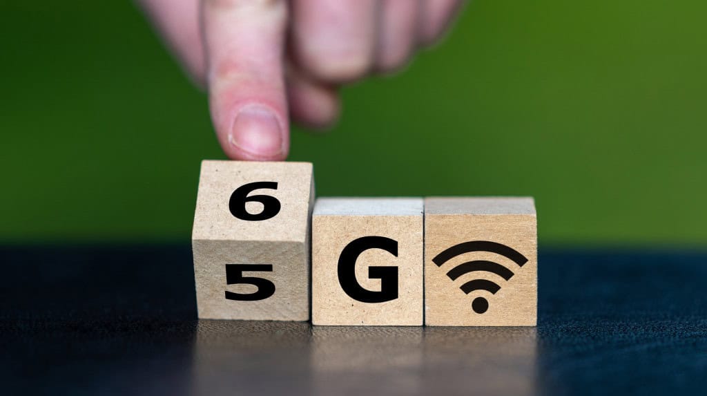 Pequenos blocos de madeira com o símbolo de internet e uma mão trocando o primeiro bloco de 5 para 6, formando a palavra "6G"