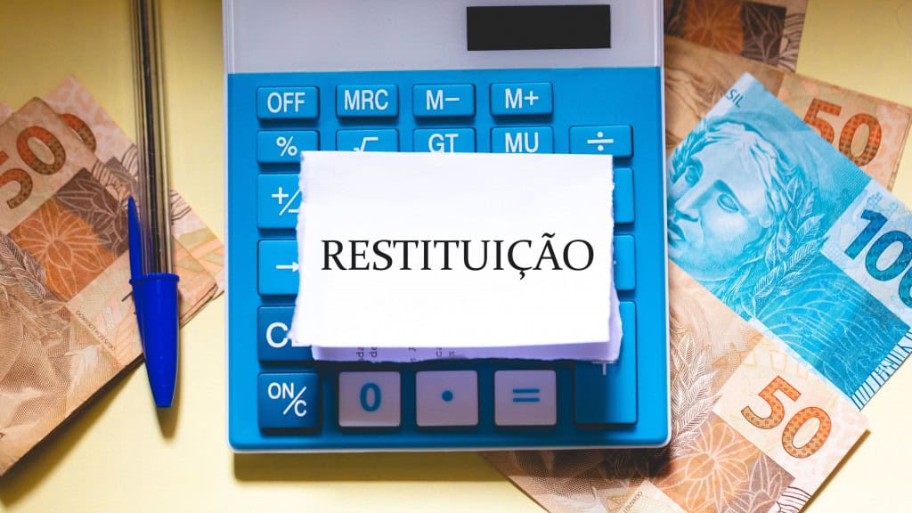 A palavra restituição em português do Brasil escrita em um pedaço de papel sobre uma calculadora. Notas de Real brasileiro na composição. Economia brasileira e imposto de renda.