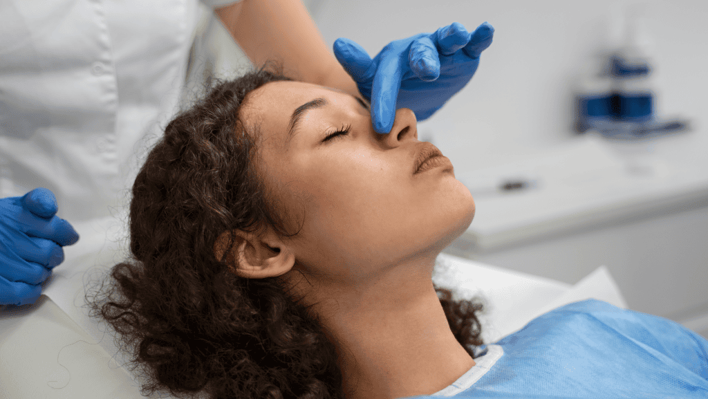 Médico com luvas azuis tocando no nariz da paciente mulher com cabelos escuros e pele clara.