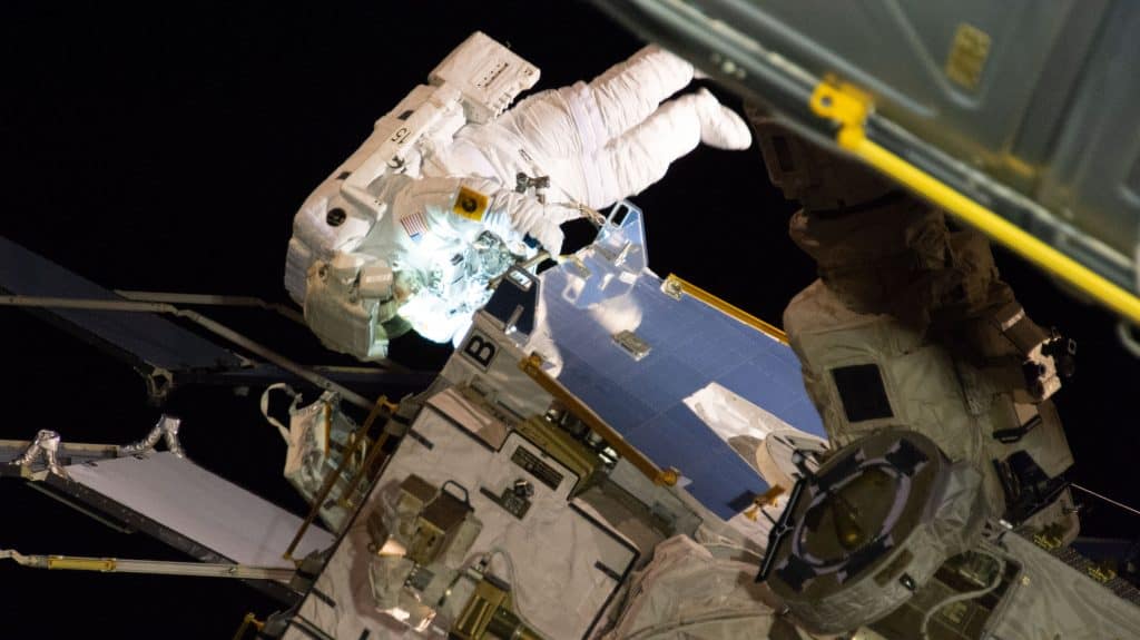 Christina Koch em traje espacial durante caminhada espacial, com a Terra parcialmente visível ao fundo enquanto ela trabalha no exterior da Estação Espacial Internacional, ajustando equipamentos no espaço aberto