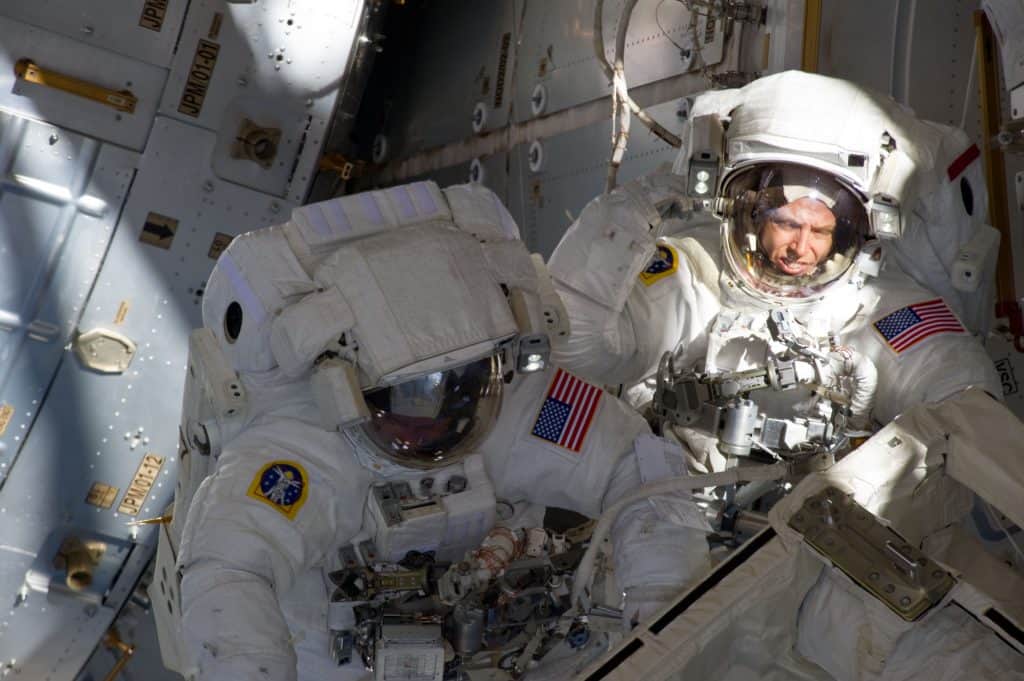 Imagem mostrando dois astronautas em trajes espaciais durante uma caminhada espacial ao redor da Estação Espacial Internacional. Estruturas da estação são visíveis, destacando os astronautas Andrew Feustel e Michael Fincke colaborando em tarefas no espaço aberto, com a Terra ao fundo.