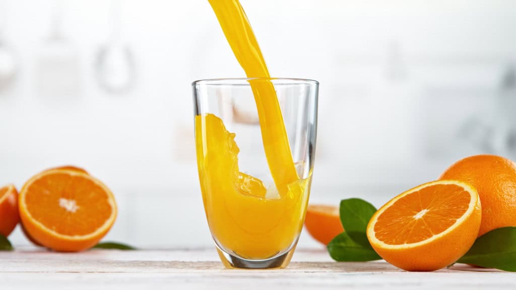 Suco de laranja sendo despejado em copo grande de vidro rodeado de laranjas cortadas