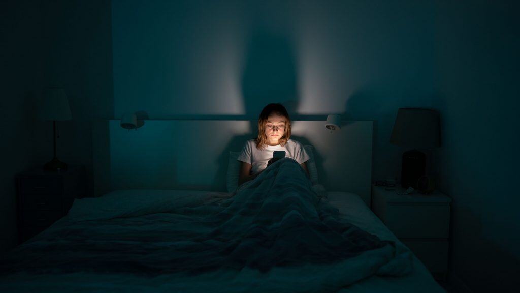 Mulher exausta deitada na cama usando o celular, sem conseguir dormir