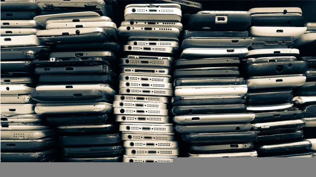 imagem mostra uma enorme pilha de celulares antigos e quebrados, uns sobre os outros, em cima de uma mesa