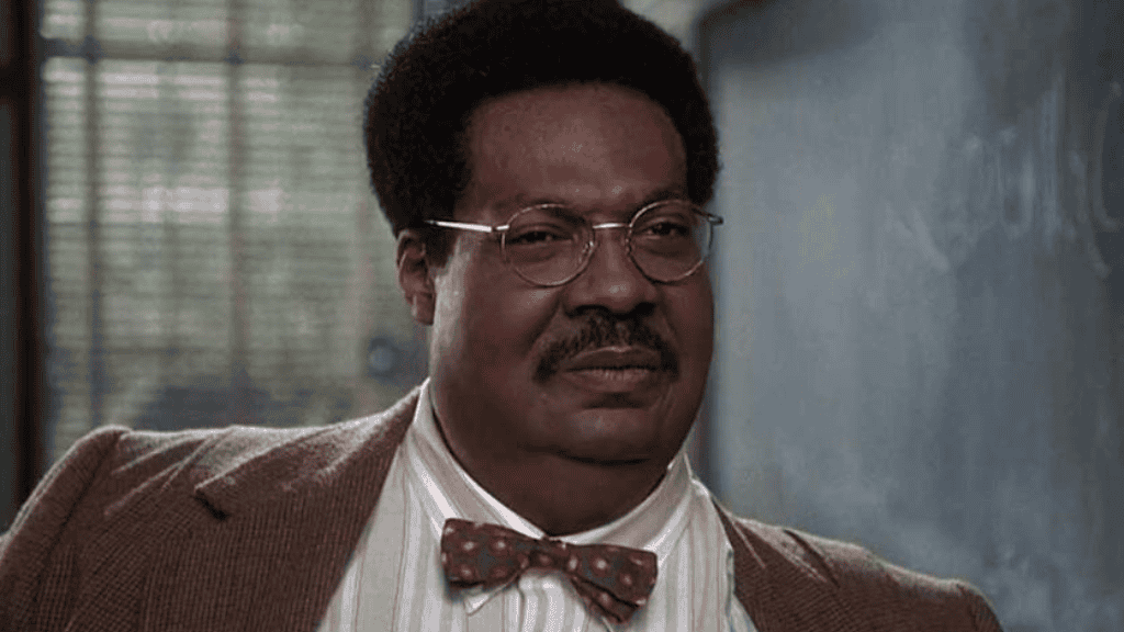 Homem negro de meia-idade, com cabelo crespo curto e volumoso, usa óculos de armação metálica, paletó marrom, camisa listrada e gravata borboleta com estampa de círculos vermelhos. Ele tem uma expressão pensativa em frente a um quadro-negro