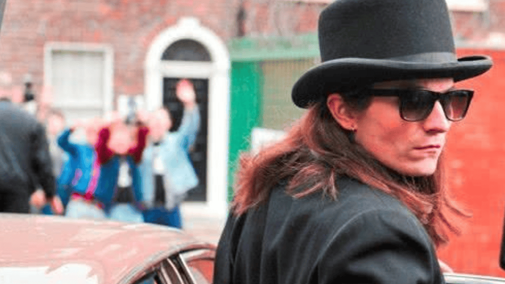 Cena do filme Matando Bono (2011). Um homem de cabelo longo veste um terno preto, usa óculos escuros e um chapéu cartola, com expressão séria. Ao fundo, um grupo de pessoas desfocado reage animadamente.