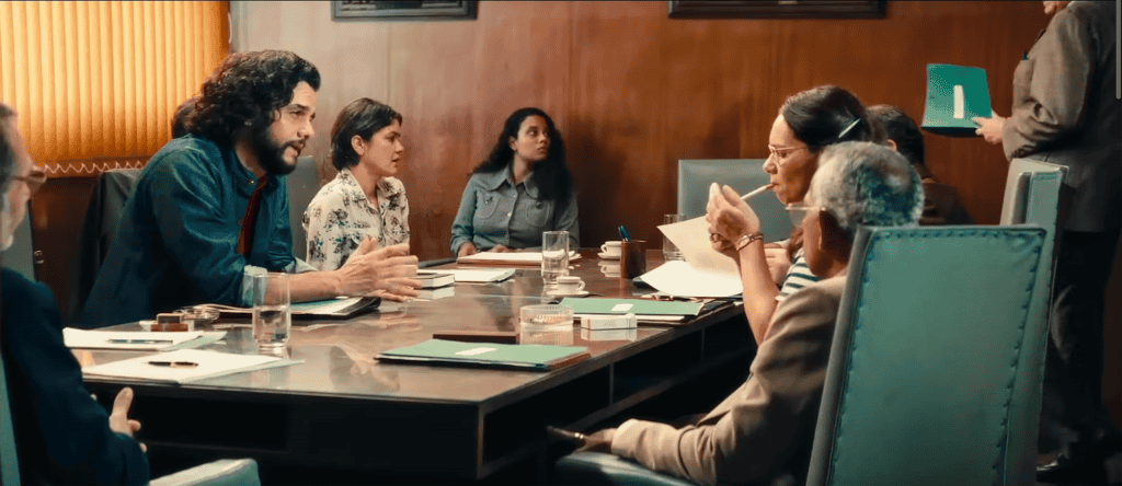 Várias pessoas sentadas ao redor de uma mesa quadrada conversando; uma delas, à direita, está acendendo um cigarro; na mesa, há vários cadernos verdes