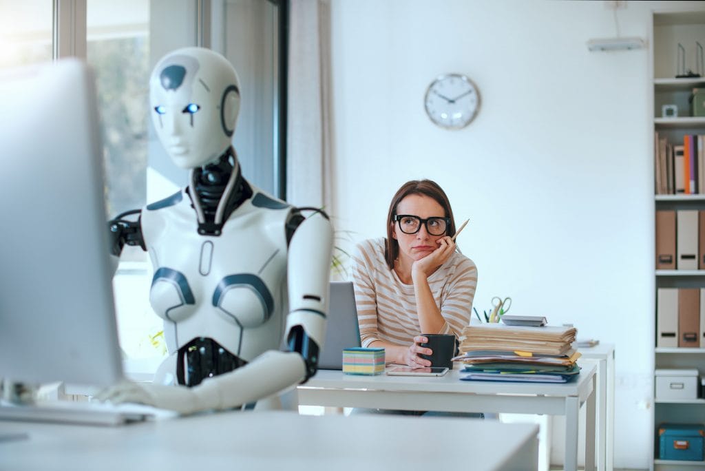 Robô sentado em frente a um computador, com uma mulher emburrada atrás olhando para ele