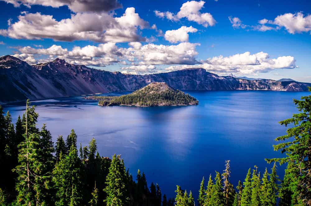 imagem mostra uma vista de cima do lago crater: a água é de um azul profundo, que combina com o azul do céu e contrasta a mata alta e verde ao redor